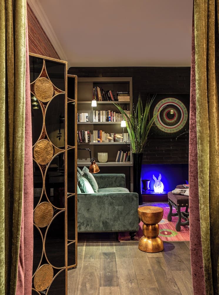 A cozy reading nook framed by elegant curtains features a plush green armchair, a bookshelf filled with books, vibrant decor including a glowing rabbit lamp and colorful rug, ideal for travelers seeking a serene and stylish retreat space.