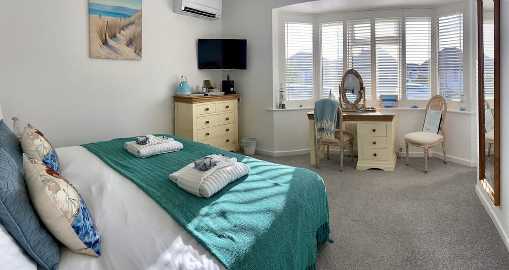 A cozy bedroom with a neatly made bed, a bay window with blinds, a vanity table, and beach-themed decor creates a serene retreat ideal for vacationers seeking relaxation.