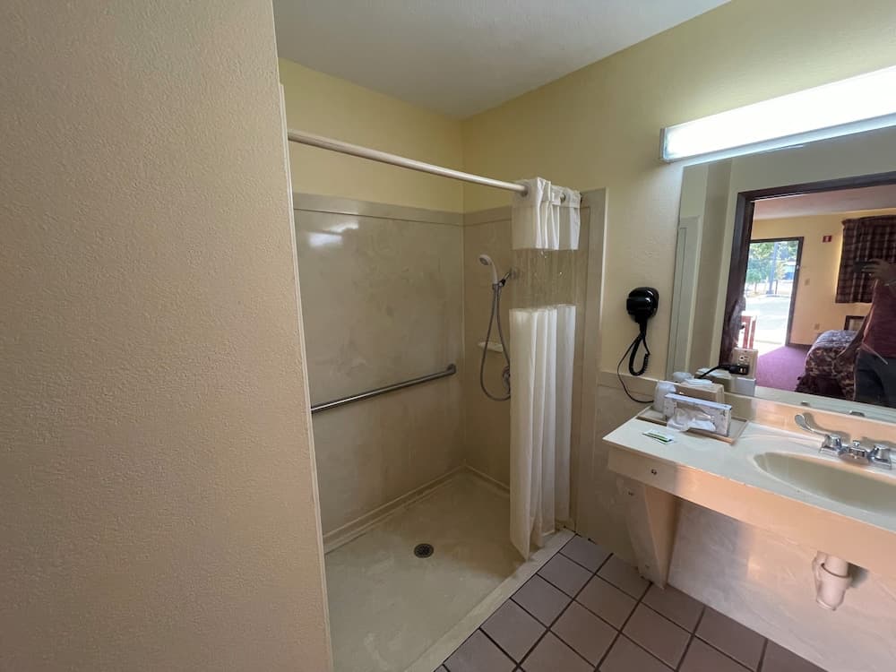 A cozy hotel bathroom features a walk-in shower with a grab bar, tiled flooring, a sink with toiletries, and a mirror reflecting a serene bedroom view, ideal for travelers seeking comfort and accessibility.