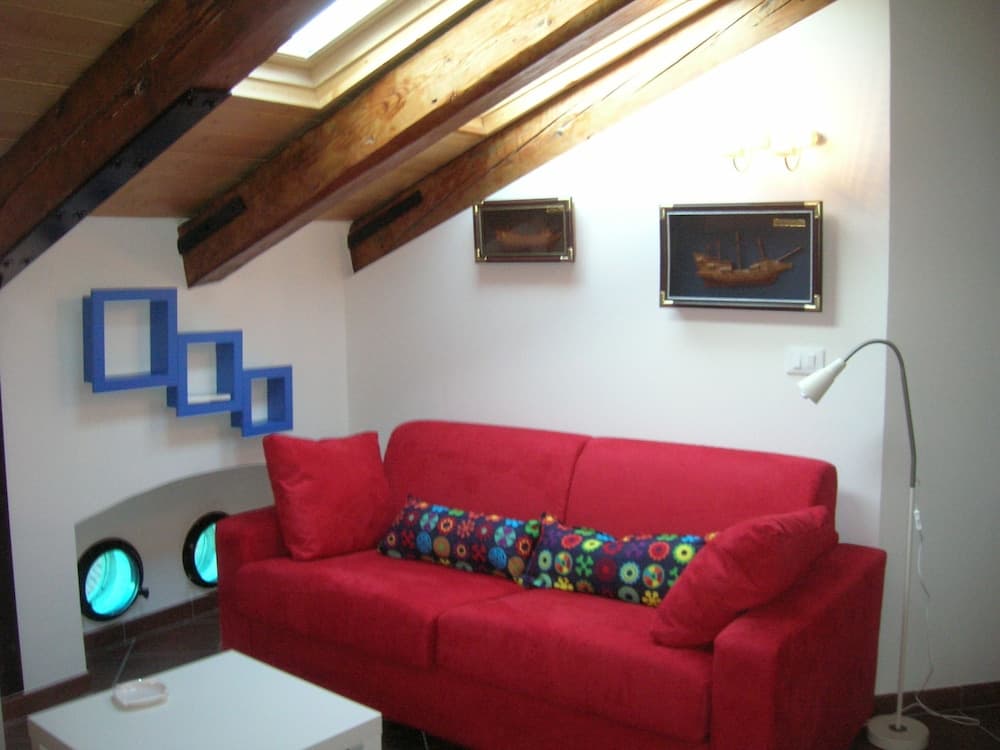 A cozy attic living space features a vibrant red sofa adorned with colorful pillows, wooden beams, nautical-themed decor including framed ship models and porthole windows, ideal for a unique vacation rental experience.