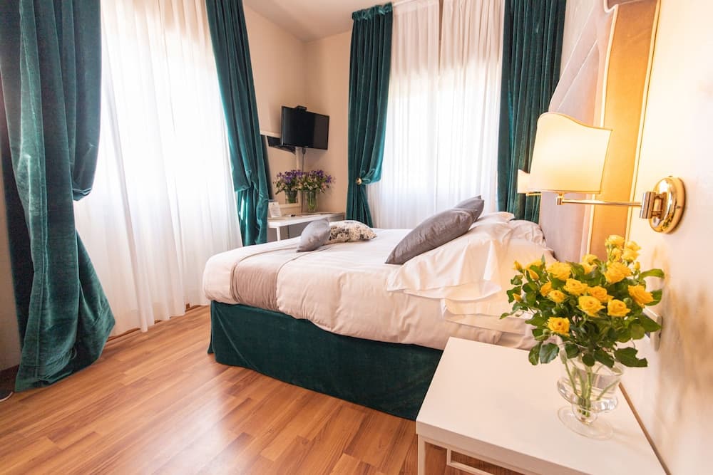 A cozy hotel room with plush teal curtains, a neatly made bed, vibrant yellow flowers on a white side table, and soft lighting creates an inviting atmosphere perfect for travelers seeking comfort and relaxation.