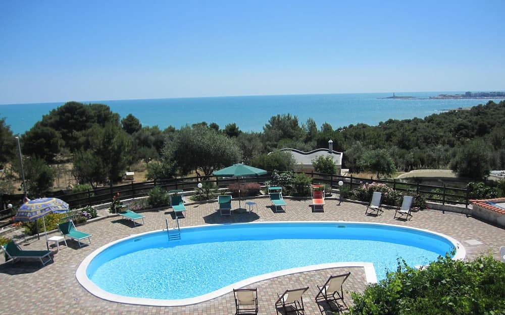A serene coastal resort features a sparkling pool surrounded by lounge chairs and umbrellas, set against lush greenery and a breathtaking ocean view, ideal for relaxing beachside vacations.