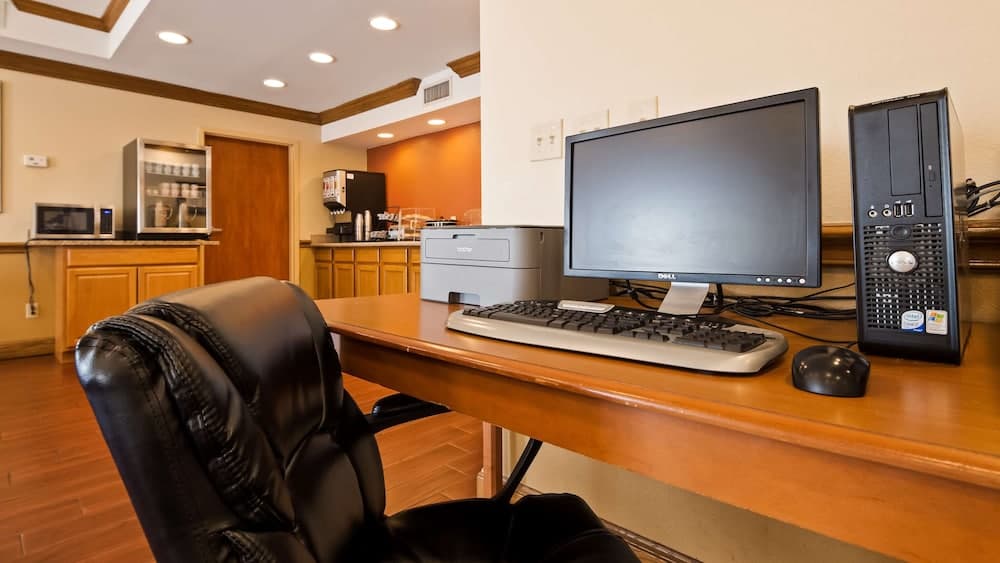 A cozy hotel business center features a wooden desk with a Dell desktop computer, printer, and leather chair, set against a warm-toned backdrop with a coffee station, ideal for travelers needing a professional workspace.