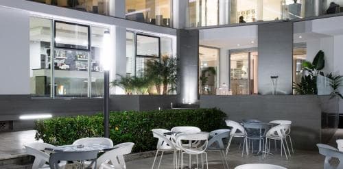 A modern urban courtyard features sleek white furniture and lush greenery under the glow of contemporary building windows, ideal for a relaxing evening retreat or business networking event.