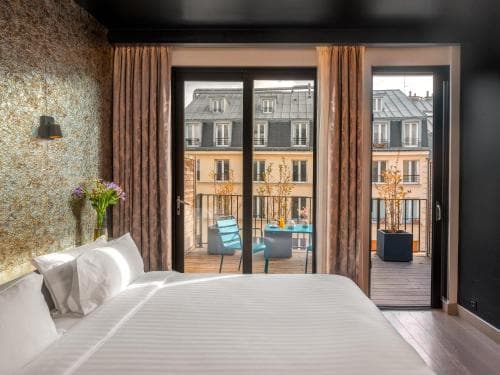 A cozy hotel room with a neatly made bed overlooks a charming balcony adorned with blue chairs and tables, perfect for travelers seeking a serene urban retreat.