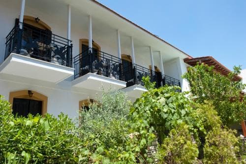 A serene Mediterranean-style hotel with white walls, balconies adorned with black railings, and lush greenery, ideal for travelers seeking a tranquil retreat in a sunny coastal destination.