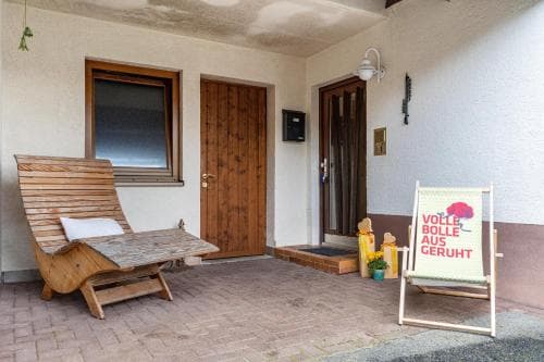 A cozy outdoor patio features a wooden lounge chair and a sign reading "VOLLE BOLLE RUHE GERUHT," inviting travelers to relax in a serene, private setting near a rustic door and window.