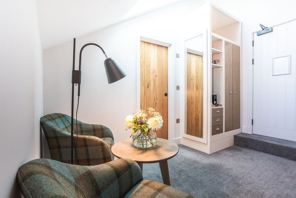 A cozy attic room features plaid armchairs, a wooden table with flowers, a modern floor lamp, and built-in storage, creating an inviting space ideal for a boutique hotel or vacation rental listing.