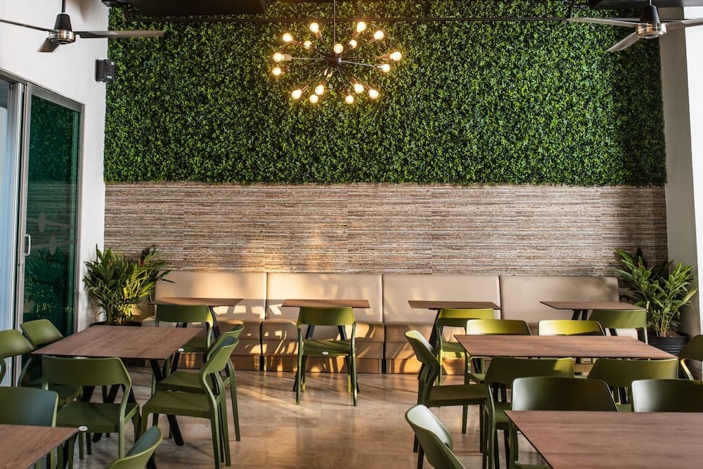 A cozy café interior features vibrant green chairs around wooden tables, a lush living wall, and a modern chandelier, creating an inviting atmosphere ideal for travelers seeking a relaxing dining experience.