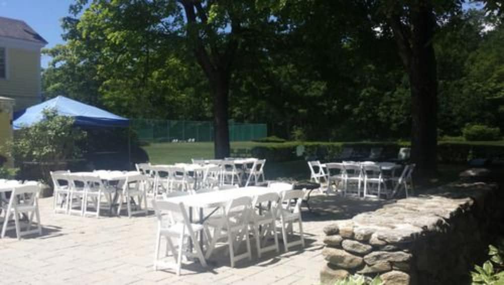 A serene outdoor patio set up with white tables and chairs under lush green trees, ideal for intimate gatherings or small events in a tranquil garden setting.