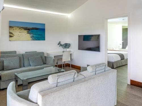 A cozy hotel suite features a modern living area with plush sofas, a glass coffee table, and beach-themed artwork, creating a serene atmosphere ideal for travelers seeking relaxation and comfort.