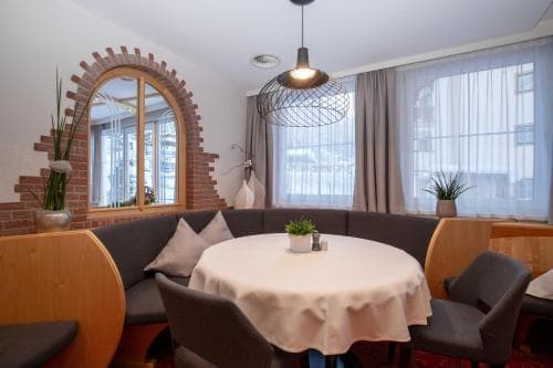 A cozy indoor dining area features a round table draped in white cloth, surrounded by modern chairs and a built-in bench under an arched brick window, creating a warm and inviting atmosphere ideal for travelers seeking a comfortable spot to relax or dine.