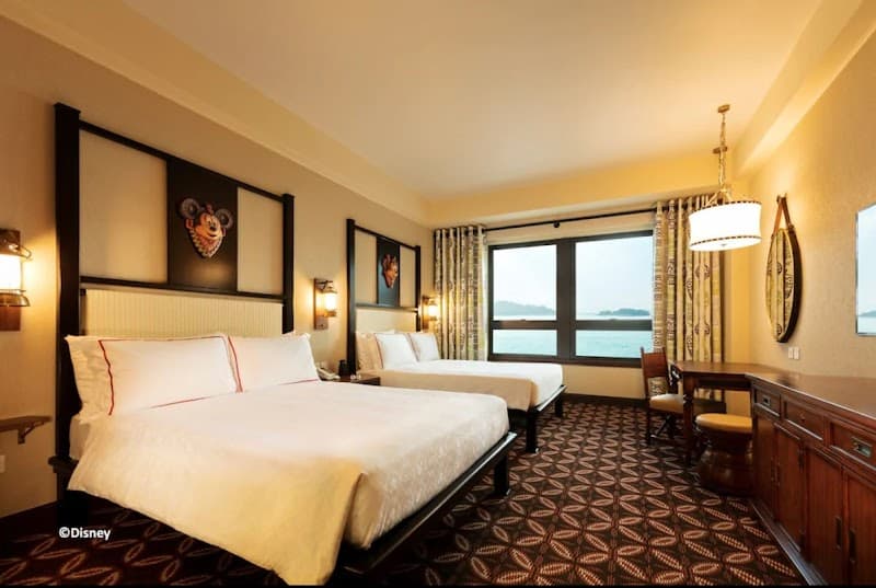 A cozy hotel room at Disney Explorers Lodge on Lantau Island, Hong Kong, features twin beds adorned with Mickey Mouse art, patterned carpeting, and a scenic view of the sea, ideal for family vacations seeking comfort and adventure.