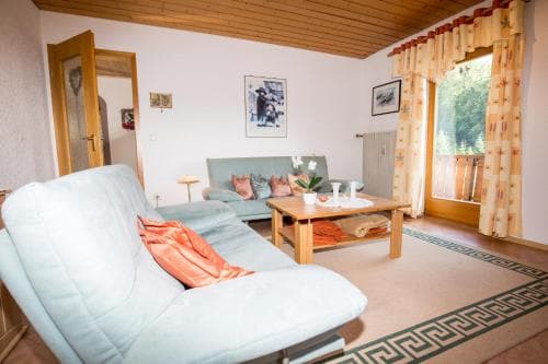 A cozy living room with light blue sofas, wooden accents, and a view of greenery through a window, ideal for a relaxing vacation rental experience.