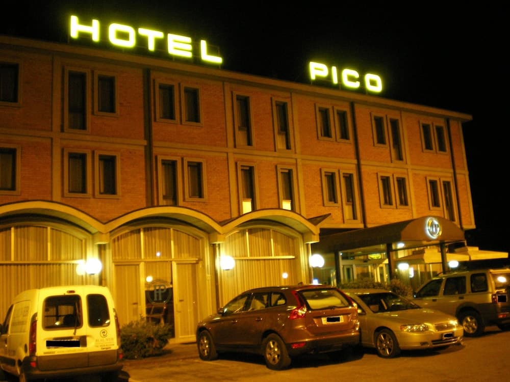 A warmly lit hotel entrance at night features a glowing "HOTEL PICO" sign, arched doorways, parked cars, and a cozy ambiance ideal for travelers seeking accommodation.