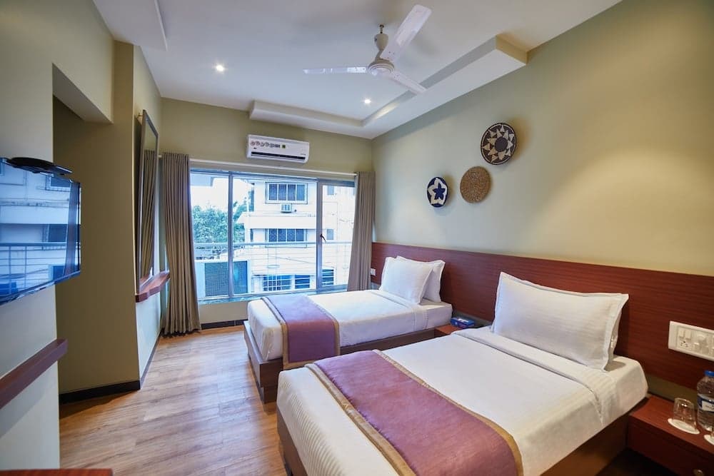 A cozy hotel room with two neatly made beds, wooden flooring, a wall-mounted TV, decorative wall art, and a balcony view of urban buildings, ideal for travelers seeking comfort and convenience.