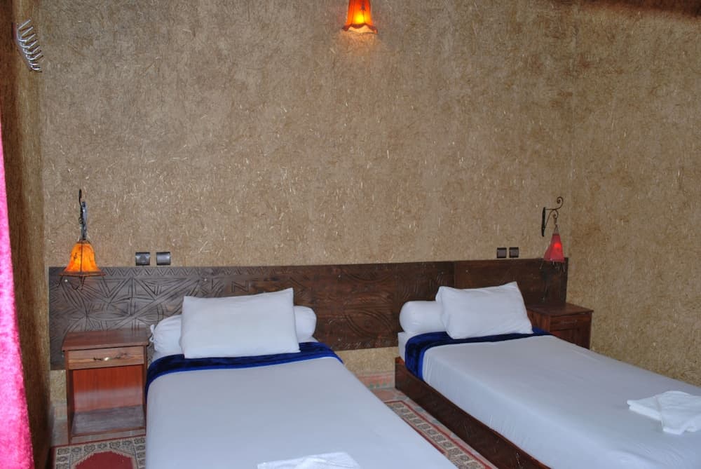 A cozy hotel room with rustic textured walls, two neatly made beds adorned with white linens and blue accents, warm lantern-style wall lamps, and a serene ambiance ideal for travelers seeking relaxation.