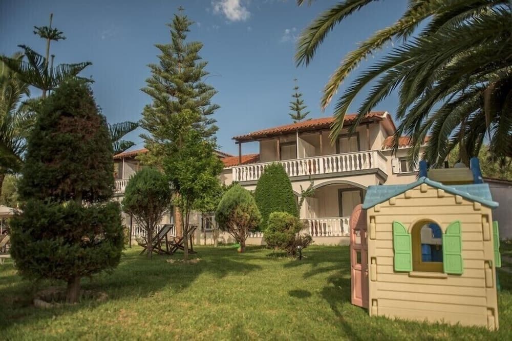 A serene family-friendly villa garden features lush greenery, palm trees, a charming playhouse, and a cozy two-story house under a clear blue sky, ideal for a relaxing vacation retreat.