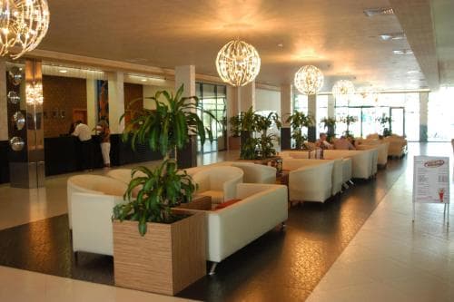 A modern hotel lobby features sleek white sofas, vibrant green plants, and spherical chandeliers, creating a welcoming atmosphere ideal for travelers seeking relaxation and comfort.