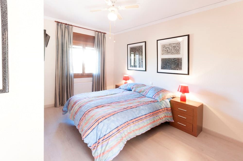 A cozy bedroom with a neatly made bed, wooden nightstands topped with red lamps, framed artwork on the walls, and soft curtains framing a window, ideal for travelers seeking a comfortable and inviting accommodation.