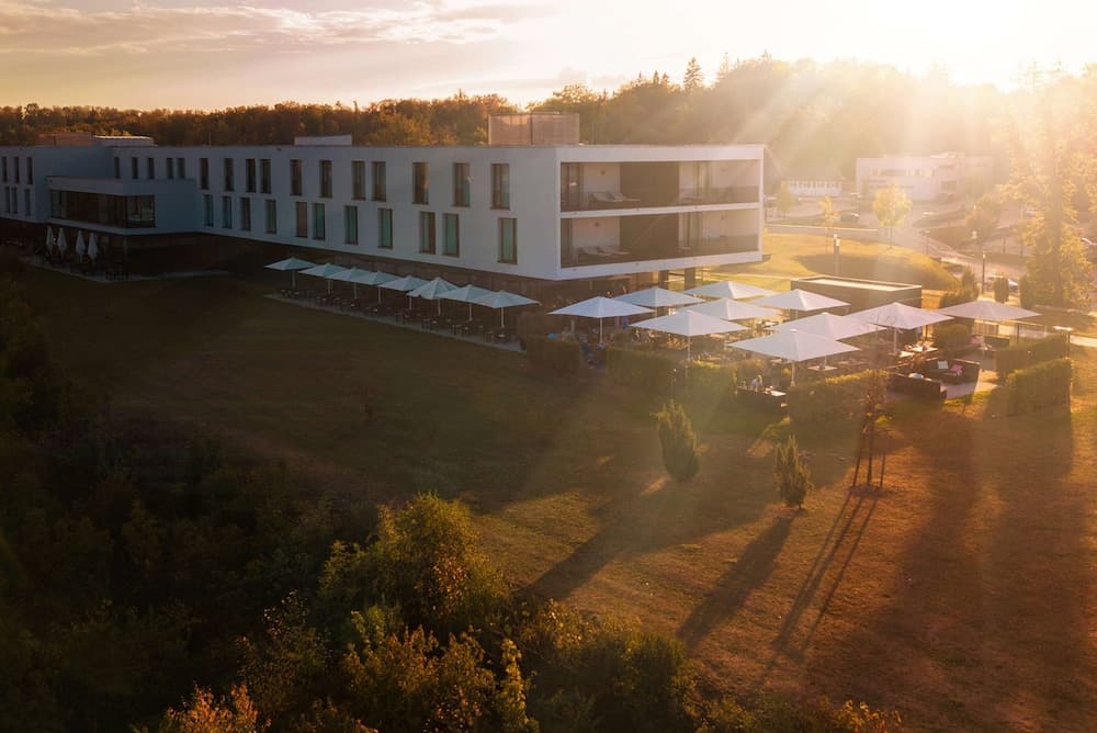 A serene sunset bathes a modern hotel surrounded by lush greenery, featuring an outdoor seating area with white umbrellas, ideal for travelers seeking relaxation and scenic views.