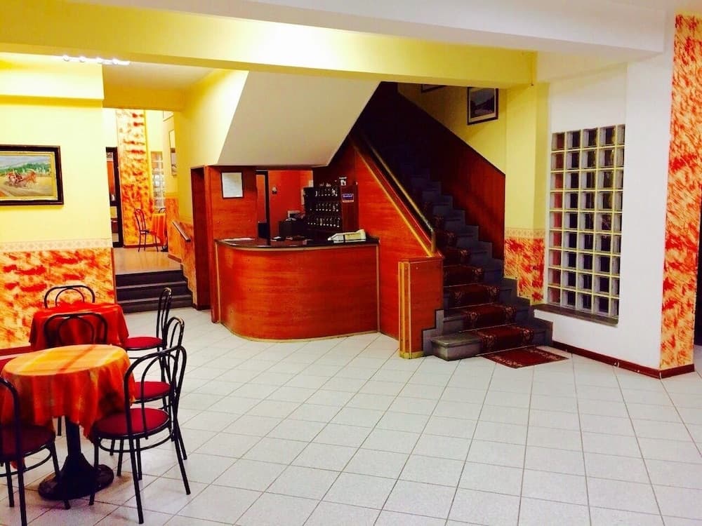 A cozy hotel lobby with warm yellow walls, tiled floors, red-clothed tables, a wooden reception desk, and a staircase leading upstairs, ideal for travelers seeking a welcoming atmosphere.