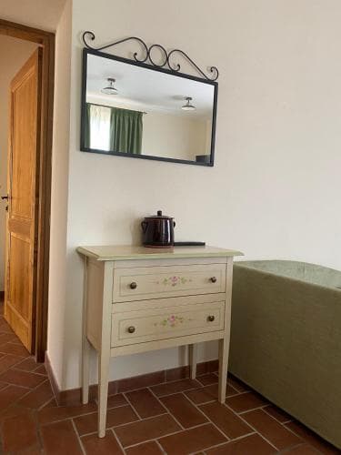 A cozy hotel room corner features a charming wooden dresser adorned with floral details, topped by a decorative teapot and framed by a wrought iron mirror, set against terracotta flooring and a soft green bedspread, ideal for travelers seeking a quaint and inviting accommodation.