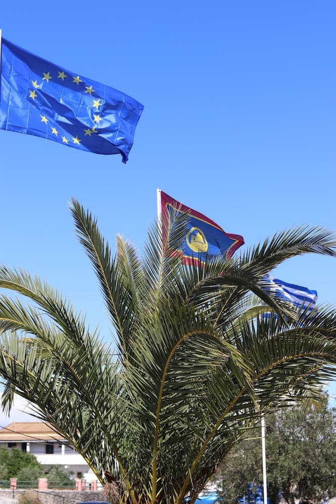 A vibrant tropical scene featuring palm fronds and fluttering European Union and Greek flags against a clear blue sky, evoking a serene seaside resort atmosphere ideal for cultural and leisure travel.