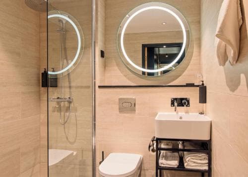 A modern bathroom with sleek tiled walls, a circular mirror with LED lighting, and a glass-enclosed shower creates a serene and luxurious atmosphere, ideal for travelers seeking comfort and style in their accommodations.
