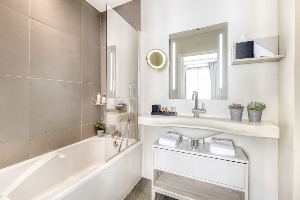 A modern, serene bathroom features a sleek bathtub with a glass shower screen, a well-lit vanity with neatly folded towels and decorative plants, ideal for travelers seeking comfort and style in their accommodations.
