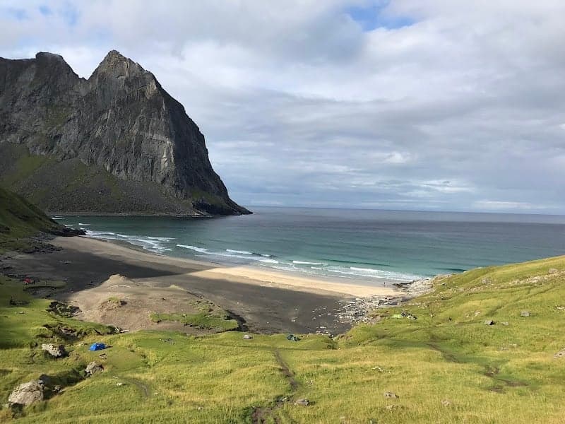 A serene Kvalvika Beach in Norway features a stunning black sand shore, turquoise waters, and a dramatic rocky cliff backdrop, ideal for tranquil camping and nature photography.