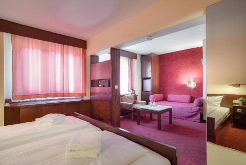 A cozy hotel suite features a neatly made bed, vibrant pink decor, and a comfortable living area, ideal for travelers seeking a relaxing and stylish accommodation experience.