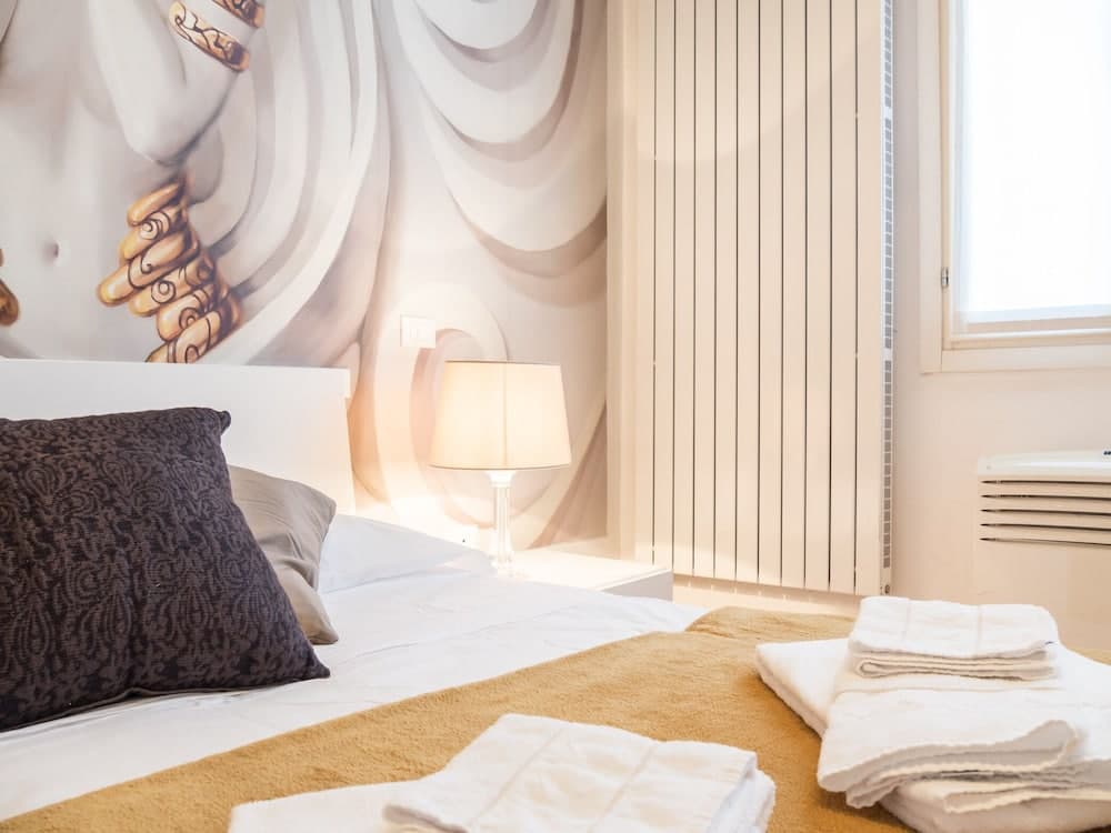 A cozy hotel room features a neatly made bed with plush pillows, a striking mural of draped fabric on the wall, soft lighting from a bedside lamp, and folded towels on the bed, ideal for travelers seeking comfort and artistry in their accommodations.