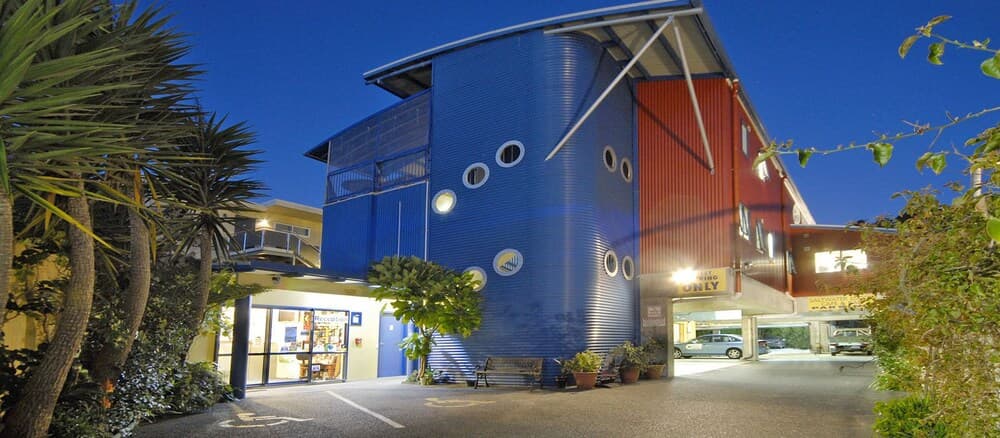A vibrant blue and red building with circular windows stands illuminated at dusk, surrounded by lush greenery, creating an inviting atmosphere ideal for travelers seeking unique accommodations or exploring urban architecture.