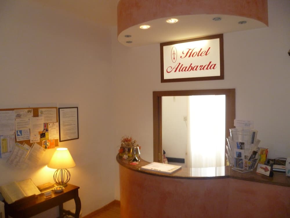 A warmly lit hotel reception area features a curved desk with brochures and a bell, a cozy lamp on a side table, and a welcoming sign above the entrance, ideal for travelers seeking comfortable accommodations.