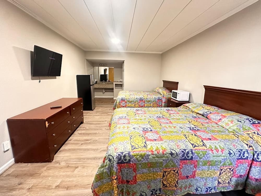 A cozy motel room features two colorful patterned beds, a wooden dresser, a mounted TV, and a small kitchenette in the background, ideal for travelers seeking comfort and convenience.