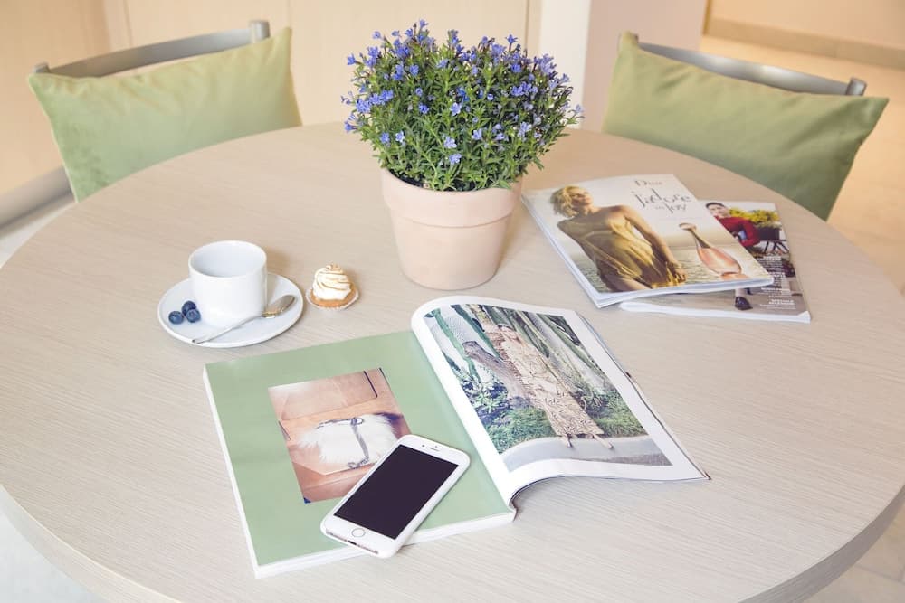 A cozy café table set with magazines, a smartphone, a cup of coffee, a small dessert, and a potted plant, evokes a relaxed and stylish atmosphere perfect for leisurely browsing or a quiet work session.