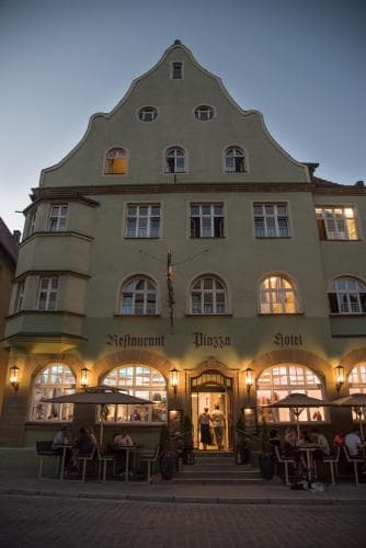 A charming European town square at dusk features a historic building housing a restaurant and hotel, with outdoor seating bustling with guests, ideal for travelers seeking cozy dining and accommodation in a picturesque setting.