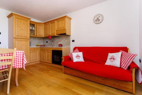 A cozy apartment kitchenette with wooden cabinets, a red sofa adorned with checkered pillows, and a dining table set with a matching tablecloth, ideal for travelers seeking a warm and inviting self-catering stay.