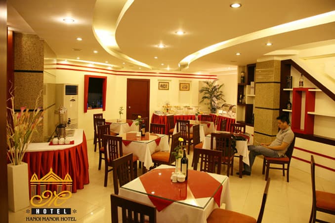 A warmly lit hotel dining area features red-accented tables, wooden chairs, and decorative plants, creating an inviting atmosphere ideal for travelers seeking a cozy meal spot.