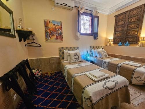 A cozy, warmly lit hotel room features two neatly made beds adorned with patterned linens, a vibrant blue carpet, ornate wooden furniture, and cultural artwork, ideal for travelers seeking a comfortable and culturally immersive stay.