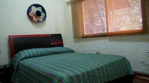 A cozy bedroom with a neatly made bed adorned in vibrant green and blue patterns, set against rustic white walls decorated with a circular wall art piece, evokes a tranquil and inviting atmosphere ideal for travelers seeking a peaceful retreat.