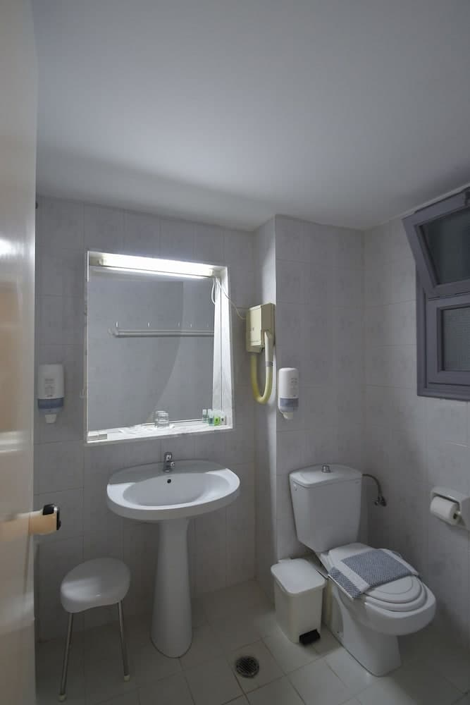 A clean, compact bathroom with white tiled walls, a pedestal sink, toilet, hairdryer, and small stool offers a functional space ideal for travelers seeking basic amenities in a hotel or guesthouse setting.