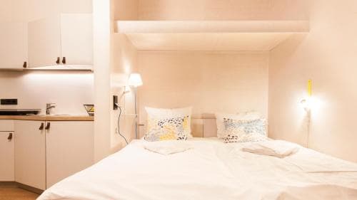 A cozy studio apartment features a neatly made bed with floral pillows, a compact kitchenette, and soft lighting, creating an inviting atmosphere ideal for solo travelers seeking comfort and convenience.