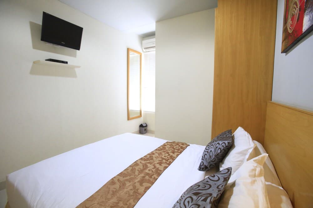 A cozy hotel room features a neatly made bed with patterned pillows, a wall-mounted TV, a wooden headboard, and soft natural light from a window, ideal for travelers seeking comfort and relaxation.