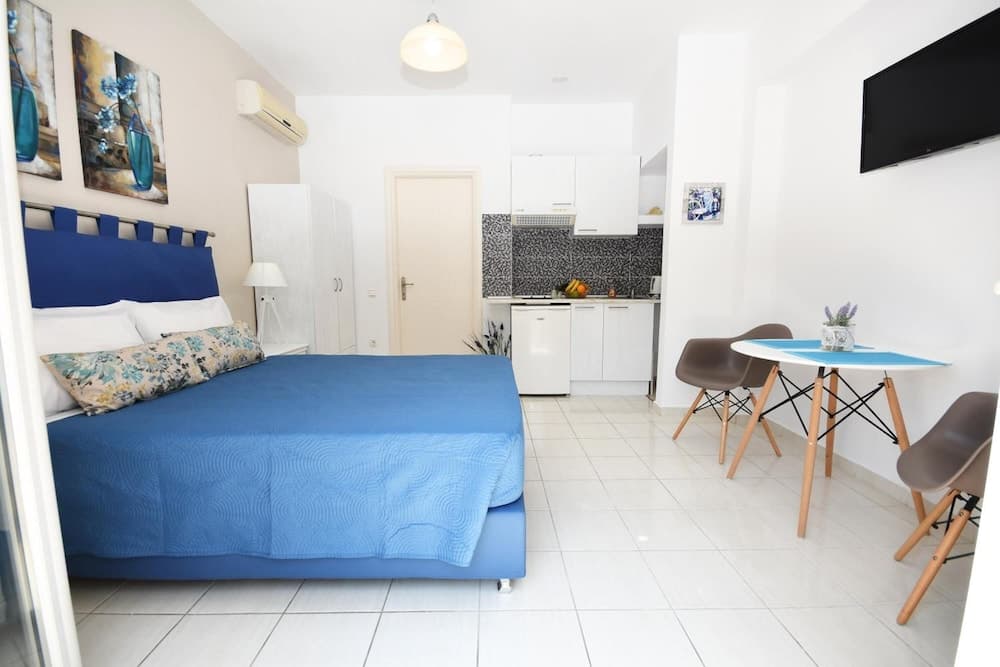 A cozy studio apartment features a blue bed, modern kitchenette, dining area with a blue table, and vibrant artwork, ideal for travelers seeking comfort and convenience in a compact space.