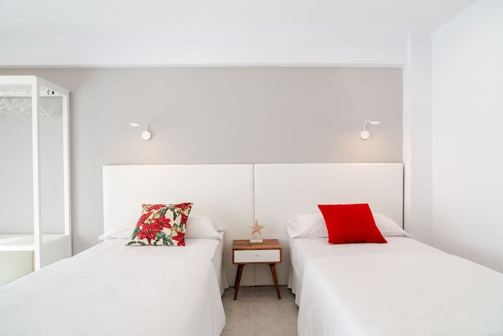A serene minimalist bedroom features two neatly made beds with vibrant pillows, a small wooden nightstand, and soft lighting, ideal for travelers seeking a cozy and tranquil accommodation.