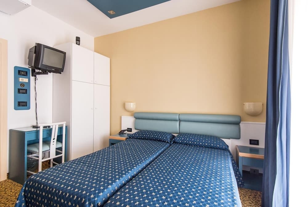 A cozy hotel room features a neatly made bed with blue floral bedding, a small desk with a CRT TV, and warm yellow walls, ideal for travelers seeking comfort and simplicity.