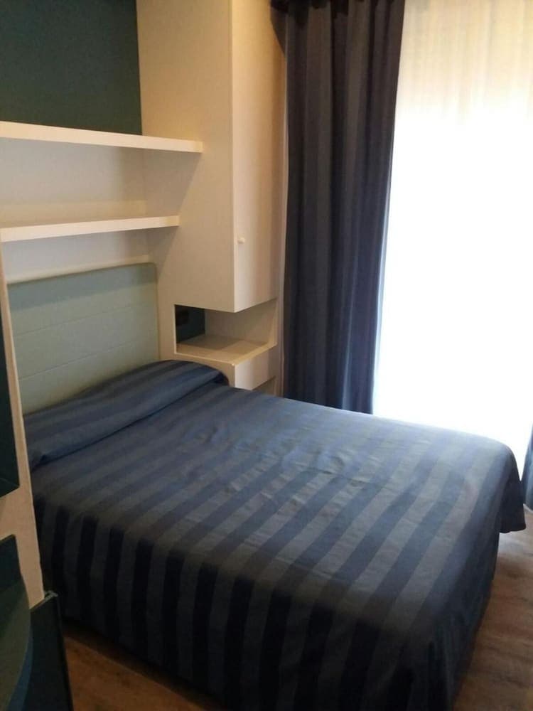 A cozy hotel room features a neatly made bed with striped blue bedding, built-in shelving, and soft natural light filtering through dark curtains, ideal for travelers seeking comfort and simplicity.