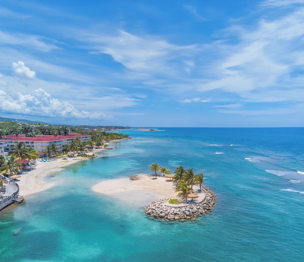 A serene tropical beach scene with crystal-clear turquoise waters, a small palm-fringed island, and resort buildings under a bright blue sky, ideal for luxury vacation planning.